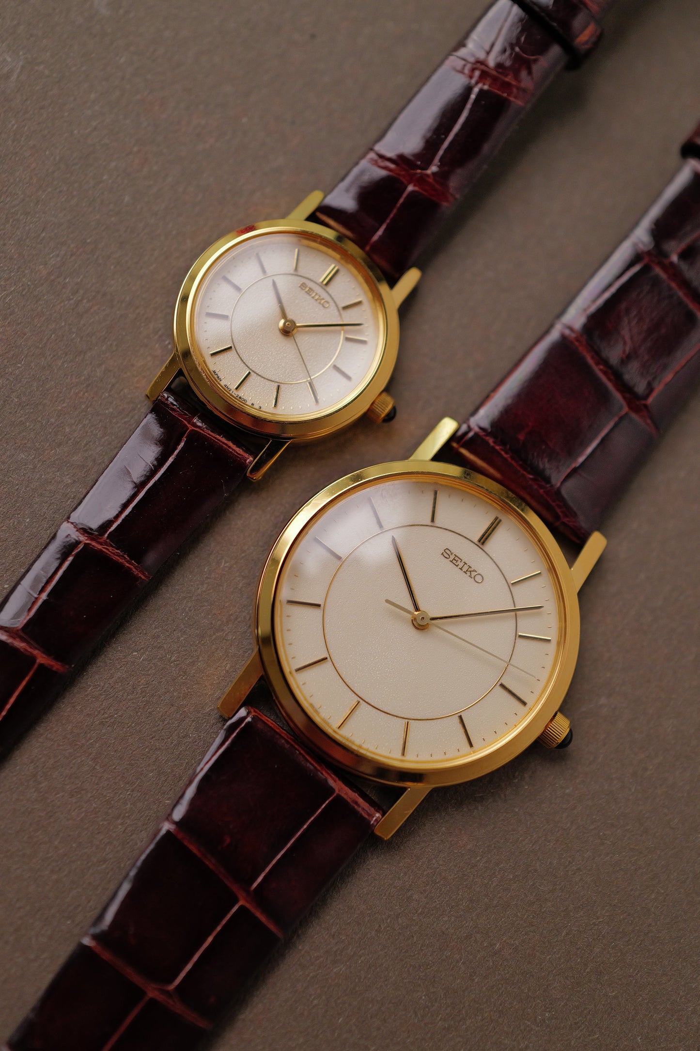 Two gold wristwatches with brown leather straps on a gray surface