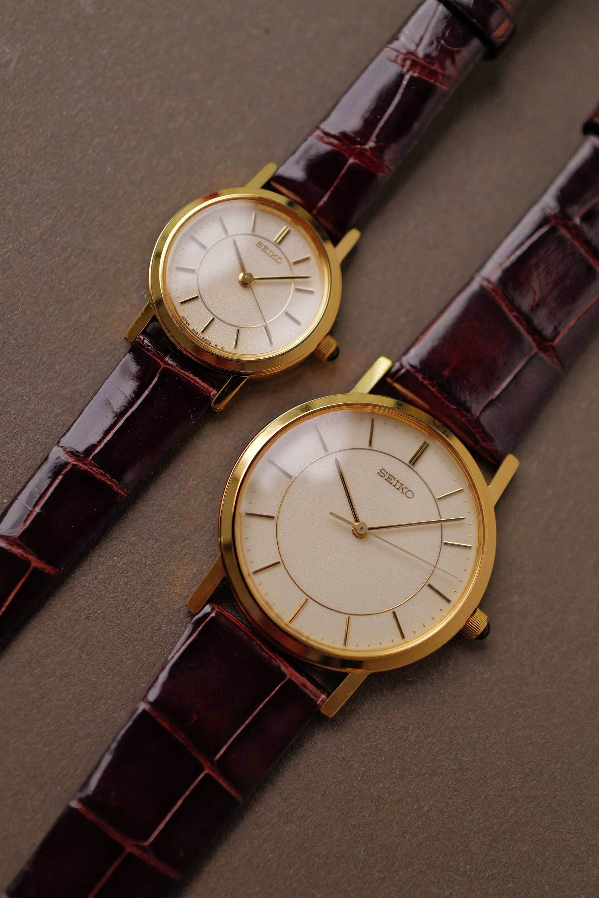 Two gold wristwatches with brown leather straps on a gray surface